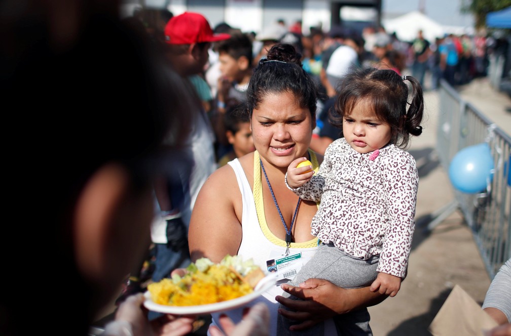 Imigrante com filha recebe neste domingo (23) prato de paella perto de abrigo temporário em Tijuana, no México — Foto Mohammed Salem Reuters